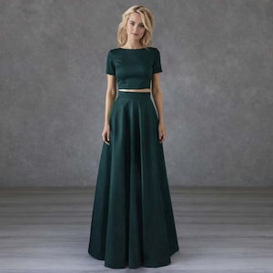 Puede incluir: Conjunto de dos piezas verde esmeralda. La parte superior es un top corto de manga corta con cuello barco. La falda es una falda larga, de línea A. El conjunto está hecho de una tela lisa y brillante. El fondo es gris liso.