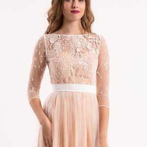 May include: A blush pink tulle dress with a white lace overlay and a white satin sash. The dress has a scoop neckline and 3/4 sleeves.
