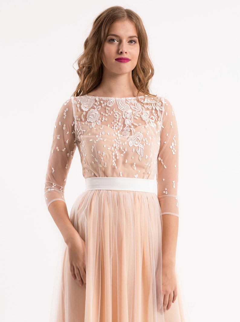 May include: A blush pink tulle dress with a white lace overlay and a white satin sash. The dress has a scoop neckline and 3/4 sleeves.