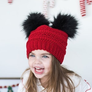 May include: A young girl wearing a red knit hat with black pom-poms and a white dress with red trim. She is smiling and looking to the right. The background is a white wall with a sign that says "Christmas" on it.