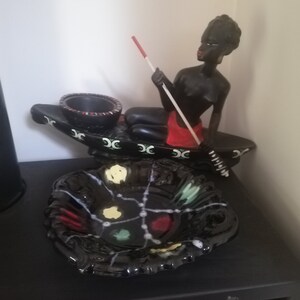May include: A black ceramic ashtray with a geometric design in red, green and white. A black figurine of a person holding a spear sits on top of the ashtray.
