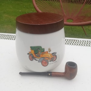 May include: White ceramic jar with a wooden lid, featuring a vintage car illustration and the text "De Dion Bouton 1903". A brown wooden pipe is lying on a white surface next to the jar.