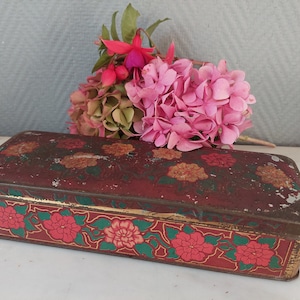 Puede incluir: Una caja de hojalata rectangular vintage con un diseño floral. La caja está decorada con flores rojas, amarillas y verdes sobre un fondo rojo oscuro. La caja tiene un borde dorado alrededor de los bordes.