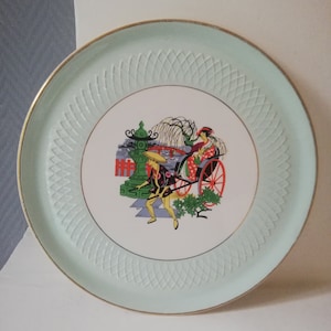 May include: A light blue ceramic plate with a gold rim features a white center with a colorful illustration of a Japanese scene. The scene depicts a person in a rickshaw being pulled by another person. The plate has a raised, textured design around the edge.