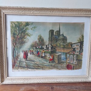 Peut inclure: Une peinture à l'aquarelle encadrée représentant une scène de rue à Paris, en France. La peinture représente la cathédrale Notre-Dame en arrière-plan, avec une rue bordée d'arbres et des gens qui marchent le long du trottoir. Il y a aussi des stands qui vendent des cartes postales et des souvenirs.