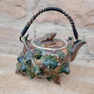 Peut inclure: Une théière en céramique artisanale avec un design texturé unique. La théière présente une palette de couleurs marron et verte avec un couvercle décoratif et une poignée enroulée de fil. La théière est conçue pour ressembler à une créature marine.