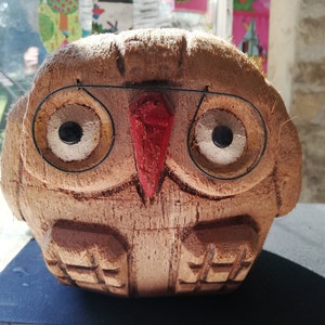 Peut inclure: Un hibou en bois sculpté dans une noix de coco avec des yeux peints et un bec rouge. Le hibou porte des lunettes à monture métallique.