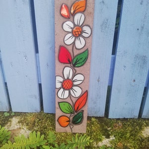 Puede incluir: Un azulejo de cerámica con un diseño pintado de una flor blanca con hojas naranjas y verdes. El azulejo es marrón y tiene una textura rugosa.