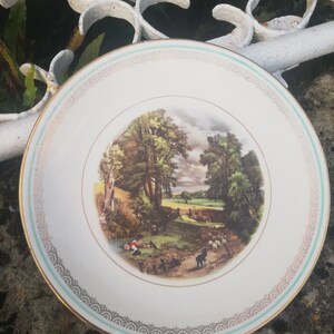 May include: A white ceramic plate with a gold rim and a green and gold trim. The plate features a vintage illustration of a rural landscape with a shepherd and sheep.