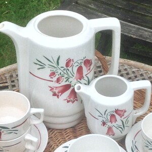 Peut inclure: Un service à thé en céramique blanche avec un motif floral. L'ensemble comprend une théière, un pot à crème, un sucrier et deux tasses et soucoupes. Le motif floral présente des fleurs rouges et vertes.