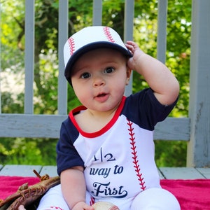 May include: A baby wearing a white and blue baseball themed outfit with a baseball cap, holding a baseball and sitting on a red blanket. The outfit has the text "1/2 way to first" on the front.