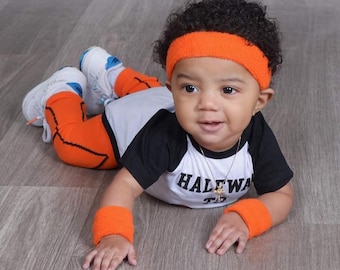May include: A baby wearing a black and white basketball jersey with the text "HALFWAY" on the front, orange and black basketball shorts, orange wristbands, and an orange headband. The baby is lying on a wooden floor.