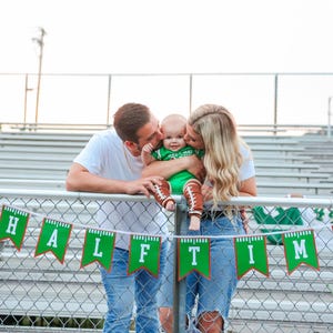 May include: A couple kisses their baby dressed in a green football jersey and brown football boots. The baby is sitting on the couple's laps. The couple is standing in front of a green and brown banner that says "HALF TIME".