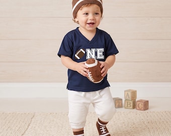 May include: A toddler in a navy blue football jersey with the word "ONE", a brown football beanie, and matching socks. The child holds a plush football. The outfit is completed with white trousers.