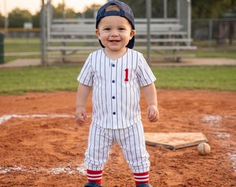 May include: A smiling toddler wearing a baseball uniform, including a navy blue cap, a white and blue striped jersey with a red number 1, matching pants, and red-striped socks. The child is standing on a baseball field.