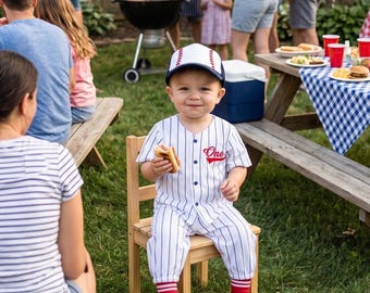 May include: A young child wearing a baseball uniform, including a cap, jersey, and pants with red socks, sits on a small wooden chair. The jersey has the word "One" on it. The child is holding a hot dog. A picnic and barbecue are in the background.