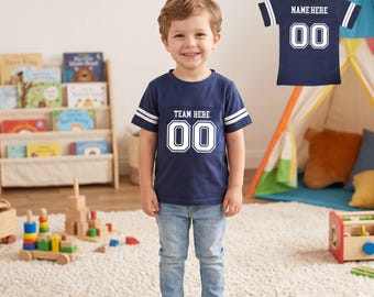 May include: A young child wearing a navy blue football-style jersey with white stripes on the sleeves. The jersey has the text "TEAM HERE" and the number "00" in white. A second jersey with "NAME HERE" and "00" is also shown.