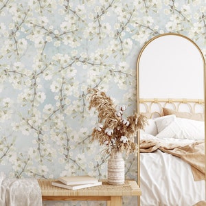 Może przedstawiać: Wnętrze sypialni z białym ł&oacute;żkiem, brązowym kocem i złotym lustrem. Ściana jest pokryta tapetą kwiatową z białymi kwiatami na jasnoniebieskim tle. Drewniana ławka z białym kocem stoi przed ścianą. Wazon z suszonymi kwiatami stoi na ławce.