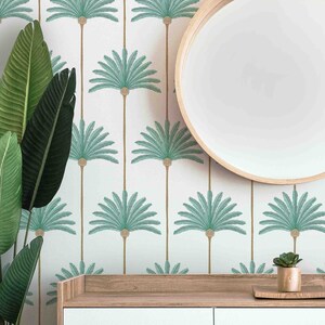 May include: A white wall with a repeating pattern of teal palm trees. A round wooden mirror hangs on the wall above a white dresser with two drawers. A small potted plant sits on the dresser.