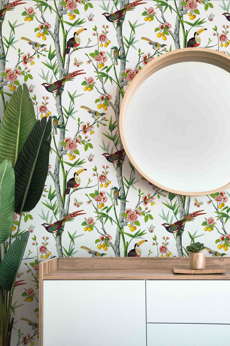 May include: White wallpaper with a repeating pattern of green bamboo branches, pink flowers, yellow fruit, and colourful birds. A round wooden mirror hangs on the wall above a white dresser with two drawers.