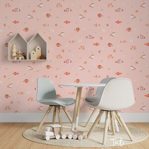 May include: A pink wallpaper with a repeating pattern of orange and white fish, starfish, and coral. Two wooden chairs and a table are in front of the wallpaper. The table is white and the chairs are light grey. There is a wooden toy train on the floor in front of the chairs.