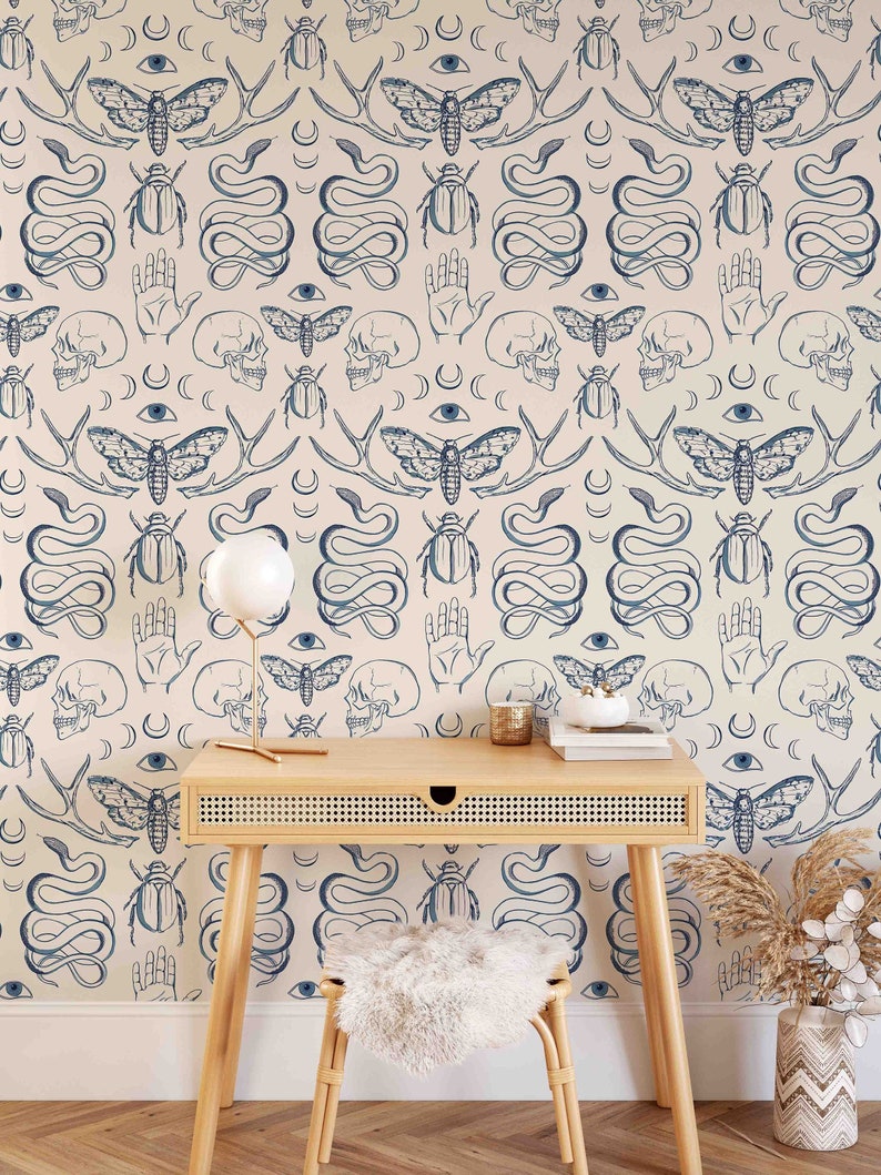 Op de afbeelding: Een lichtbeige behang met een herhalend patroon van blauwe illustraties. Het patroon omvat schedels, nachtvlinders, kevers, slangen, handen, ogen en halve manen. Een houten bureau met een lade en een witte, harige kruk staan voor het behang.