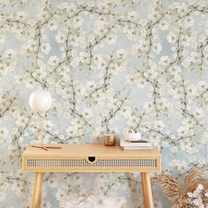 Może przedstawiać: Jasnoniebieska tapeta z białym wzorem kwiatowym. Przed tapetą stoi drewniane biurko z plecioną szufladą. Przed biurkiem stoi biały, puszysty stołek. Na biurku stoi biała lampa ze złotą podstawą. Obok biurka na podłodze stoi wazon z suszoną trawą pampasową.