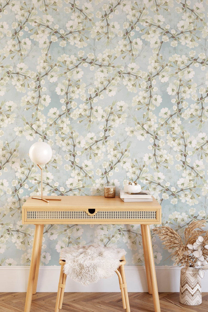 Może przedstawiać: Jasnoniebieska tapeta z białym wzorem kwiatowym. Przed tapetą stoi drewniane biurko z plecioną szufladą. Przed biurkiem stoi biały, puszysty stołek. Na biurku stoi biała lampa ze złotą podstawą. Obok biurka na podłodze stoi wazon z suszoną trawą pampasową.
