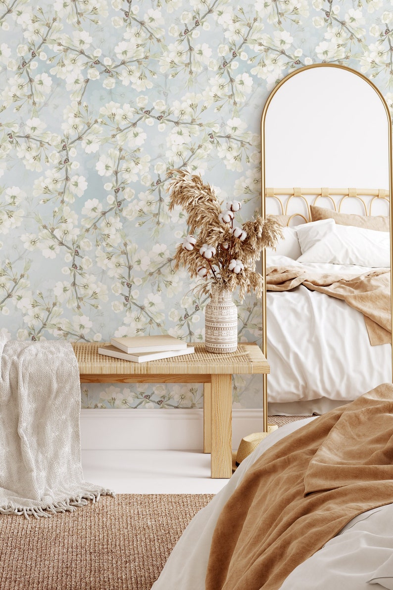 Może przedstawiać: Wnętrze sypialni z białym ł&oacute;żkiem, brązowym kocem i złotym lustrem. Ściana jest pokryta tapetą kwiatową z białymi kwiatami na jasnoniebieskim tle. Drewniana ławka z białym kocem stoi przed ścianą. Wazon z suszonymi kwiatami stoi na ławce.