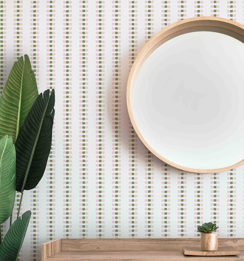 Op de afbeelding: Interieur met een witte muur versierd met verticale rijen roze en groene vierkanten. Een ronde houten spiegel hangt aan de muur. Groene planten staan links en een houten plank met een kleine plant staat beneden.