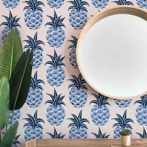 May include: A pink wallpaper with a repeating pattern of blue pineapples. The wallpaper is shown on a wall with a round mirror and a small wooden table with a potted plant.