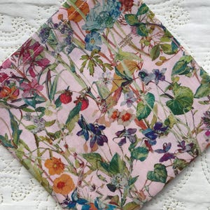 Può includere: Un tovagliolo quadrato in tessuto con un motivo floreale su sfondo rosa chiaro. Il design presenta una varietà di fiori colorati, tra cui fiori blu, arancioni e viola, insieme a foglie verdi. Il tovagliolo ha un bordo cucito bianco.