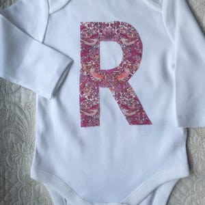 Può includere: Body da bambino bianco a maniche lunghe, con una grande lettera "R" in un motivo floreale. Il design floreale incorpora tonalità rosa, pesca e verdi. Il body ha uno scollo tondo e chiusure a scatto sul fondo.
