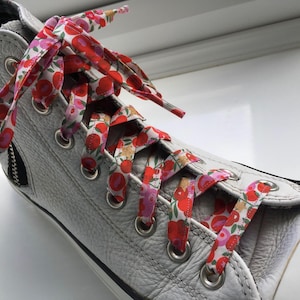 May include: Close-up of a grey high-top trainer with floral print laces. The laces are white with a pattern of red and pink flowers. The shoe has a zip on the side and silver eyelets.