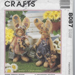 Puede incluir: Sobre de patrón de costura McCall's Crafts 8087 con dos muñecos de conejo de peluche color canela. Uno lleva un gorro floral y vestido, el otro un peto de cuadros azules. Ambos tienen parches en las orejas y sostienen letreros que dicen Sweet Spring Thyme y Fresh Flowers. Los conejos miden 41 cm de altura.