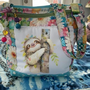 Könnte beinhalten: Eine farbenfrohe Tragetasche mit einem floralen Patchwork-Design und einer skurrilen Faultier-Illustration. Die Tasche hat einen türkisfarbenen Boden, einen grünen Reißverschluss und Perlenanhänger. Die Träger und Seiten sind mit Blumenmustern verziert.