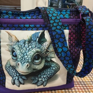 May include: A quilted shoulder bag featuring a dragon scale pattern in shades of blue, purple, and teal. The front panel displays a large, detailed graphic of a baby dragon with large eyes. A zipper closure and a beaded charm are attached.