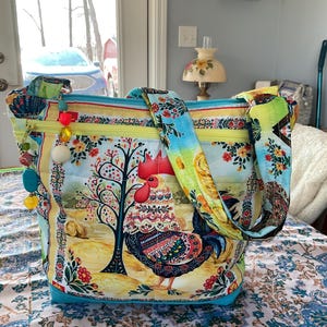 May include: A colorful tote bag featuring a rooster design. The bag has a blue base, yellow zipper, and a strap with floral and rooster patterns. Decorative beads hang from the zipper. The bag's main design includes a rooster, tree, and floral elements.