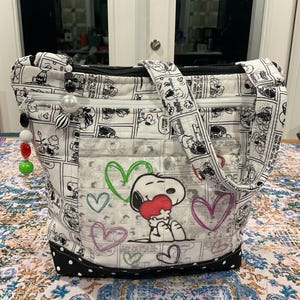 Op de afbeelding: Een gewatteerde draagtas met een zwart-wit stripverhaalpatroon. De tas heeft een Snoopy-afbeelding met een hart, met gekleurde harten eroverheen. De tas heeft een zwarte rits, handvatten en een zwarte stippelbodem.