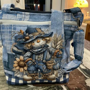 May include: A blue patchwork fabric crossbody bag featuring a central graphic of a scarecrow wearing a hat and overalls, surrounded by sunflowers and wheat. The bag has a white zipper closure, a wide shoulder strap, and a blue and white bead charm.