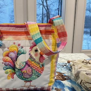 Op de afbeelding: Een kleurrijke draagtas met een grillige haanillustratie. De tas heeft een regenboog gestreept patroon, een ritssluiting en een kralenbedel. De haan heeft levendige veren in tinten blauw, groen, geel en roze. De handvatten van de tas zijn ook regenboog gestreept.