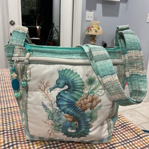 May include: A teal and white fabric tote bag with a zipper closure and a seahorse design. The bag has a long strap and is decorated with beads and charms.