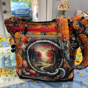 May include: A quilted tote bag with a fall-themed design. The bag features a central image of a landscape with a river, trees, and pumpkins. The bag has orange and black floral patterned straps and trim, with a red zipper.