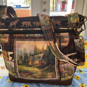 May include: A brown and black quilted tote bag with a cabin scene and bears printed on it. The bag has a brown leather bottom and a long adjustable strap.