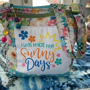 Könnte beinhalten: Blumenmuster-Tragetasche mit weißer Vordertasche. Die Tasche trägt den Text "I WAS MADE FOR SUNNY DAYS" in bunten Buchstaben. Die Tasche hat einen Reißverschluss, einen Schulterriemen und Perlenanhänger.