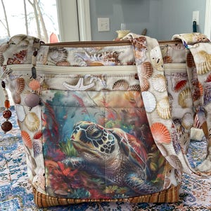 May include: A large tote bag featuring a colorful sea turtle design on the front. The bag is adorned with seashell patterns and has a brown zipper. The straps and sides are decorated with various seashells. The bag has a beaded charm.