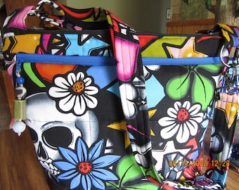 May include: A fabric shoulder bag featuring a vibrant, multicolored print of skulls, daisies, and stars on a black background. The bag includes a horizontal front zipper pocket with a blue zipper and a decorative beaded pull charm attached to the side.