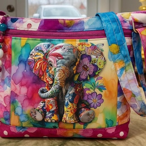 May include: A colorful tote bag featuring a whimsical elephant design. The elephant is adorned with floral patterns in shades of orange, purple, and blue. The bag has a pink polka-dot base and a blue strap, with a beaded charm.