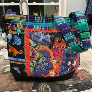 Peut inclure: Sac fourre-tout coloré avec un motif de chats. Le sac a une base noire, une fermeture éclair turquoise et une bandoulière à motifs. Le motif principal comprend des visages de chats, des motifs floraux et diverses illustrations de chats aux couleurs vives.