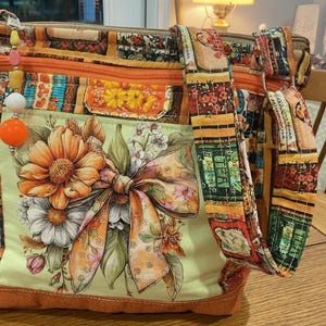 May include: A quilted fabric crossbody bag featuring a patchwork design with orange, yellow, and green floral patterns. The front panel displays a large printed bow and flower bouquet. It includes an orange zipper and a beaded charm pull.